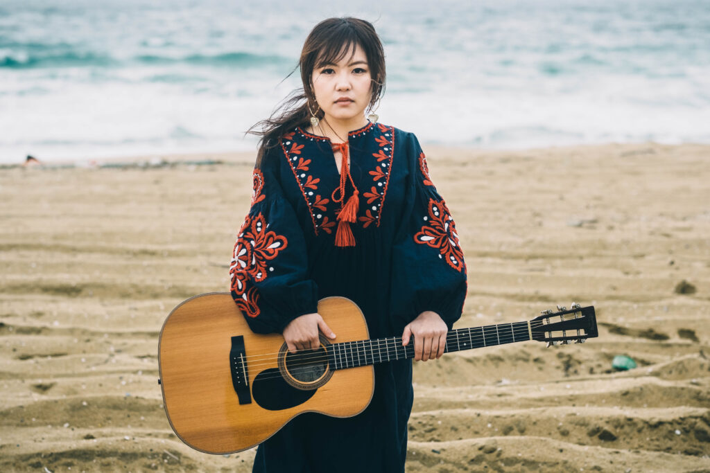 海辺でアコースティックギターを持つ松尾知華先生のアーティスト写真