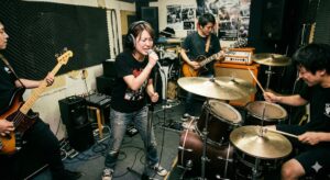 狭い音楽スタジオでドラムやギターの音圧に押され、喉を締め付けて叫ぶように歌うバンドボーカルの様子