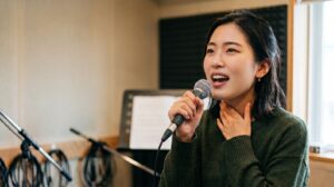 ボーカルレッスンで歌い方に悩む日本人女性。音楽教室のスタジオでマイクを持ち、喉の不調や発声の難しさを感じているリアルな表情。アイキャッチ用。